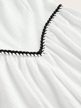 This charming white dress features delicate textured fabric with a playful black braided trim along the neckline, adding a.