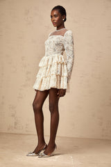Woman wearing a beige mini dress with delicate floral embroidery detail standing against a plain background