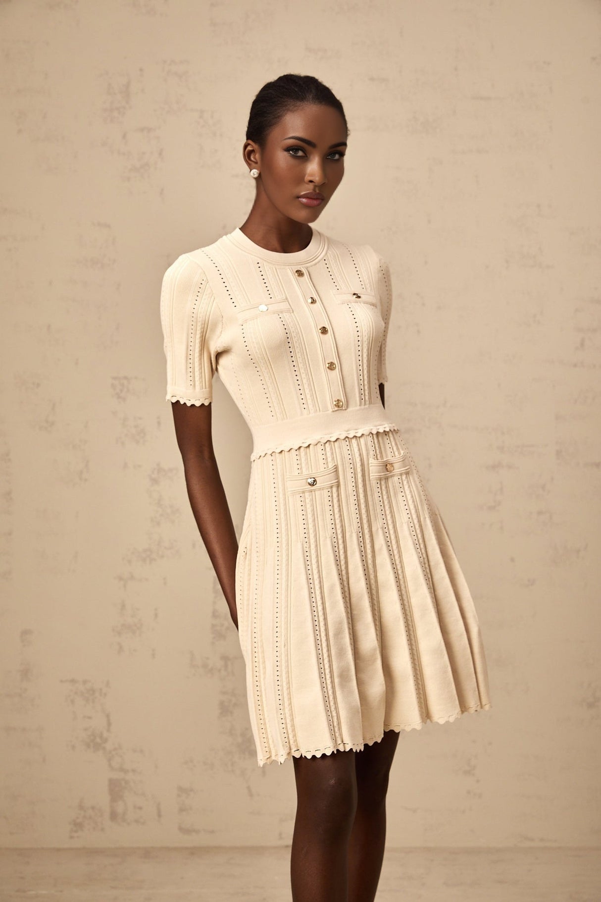 Woman wearing a beige mini dress with pleated skirt and delicate pointelle trim detail standing in a neutral setting