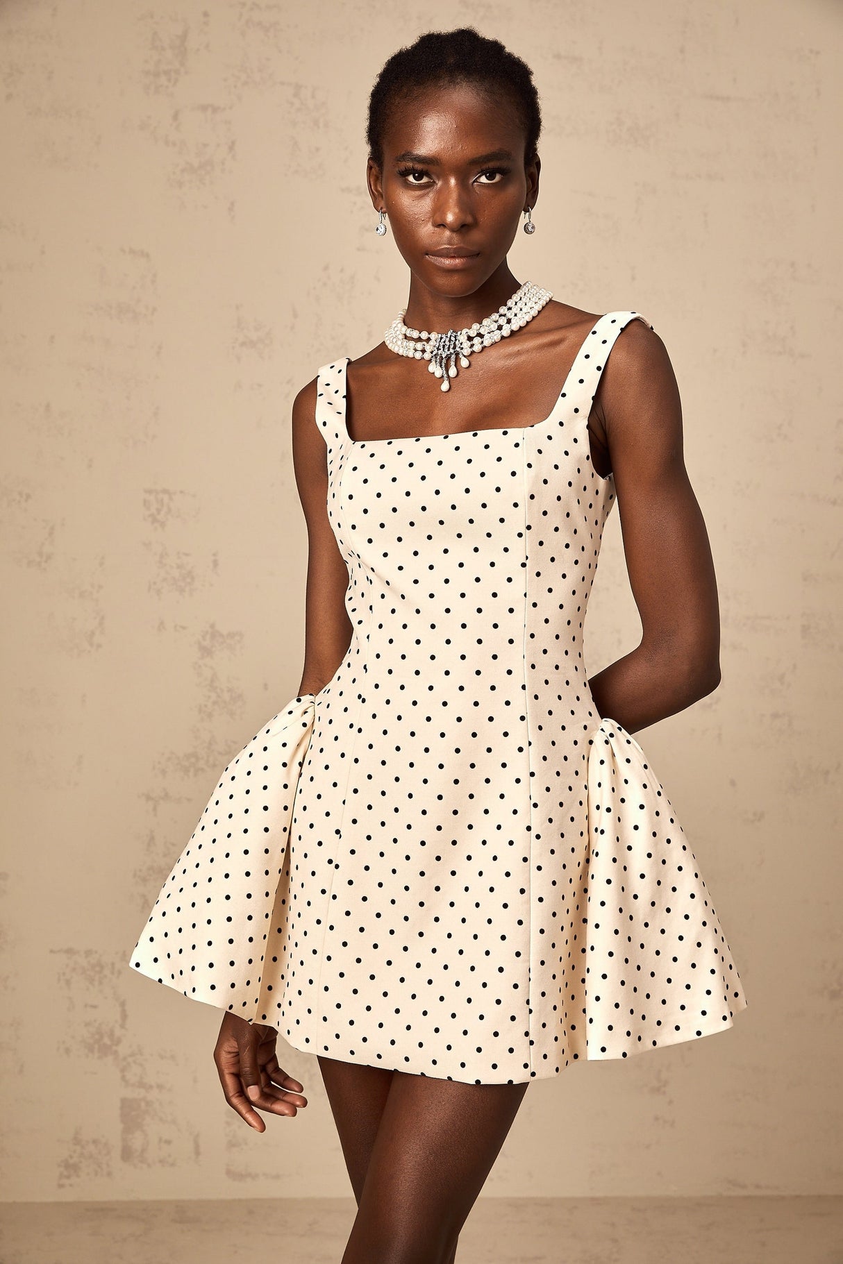 Woman wearing a beige mini dress with white polka dots and puff sleeves standing against a plain background