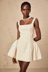 Woman wearing a beige mini dress with white polka dots and puff sleeves standing against a plain background