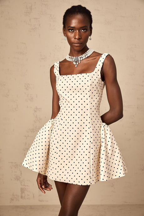Woman wearing a beige mini dress with white polka dots and puff sleeves standing against a plain background