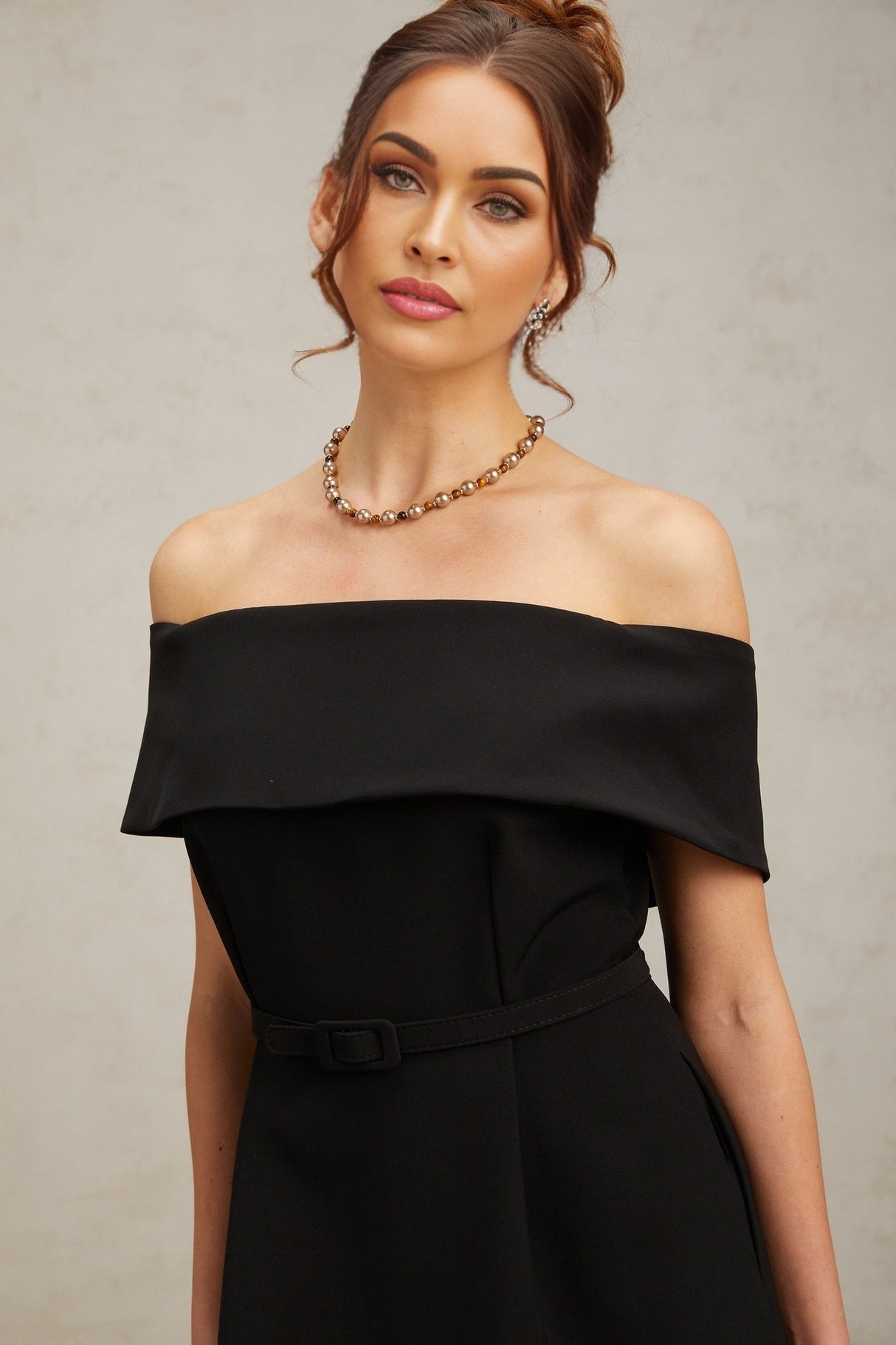 Woman wearing a black offshoulder mini dress with a belt cinched at the waist standing against a plain background