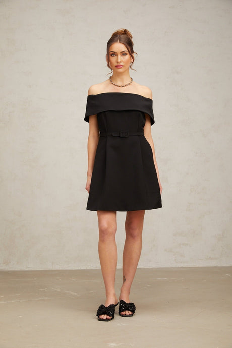 Woman wearing a black offshoulder mini dress with a belt cinching the waist standing against a plain background