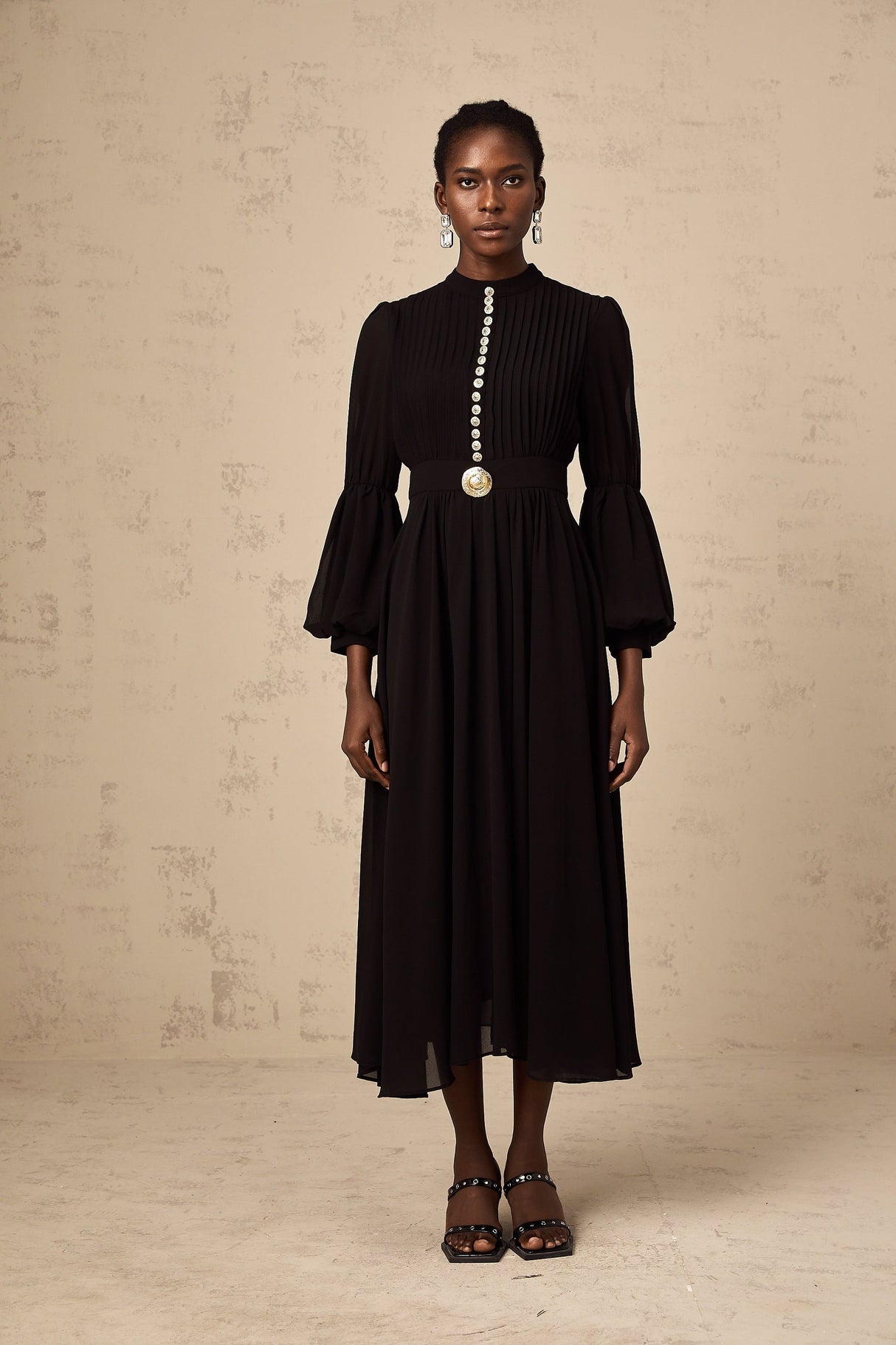 Woman wearing a black pleated midi dress with button front detail standing in a neutral pose