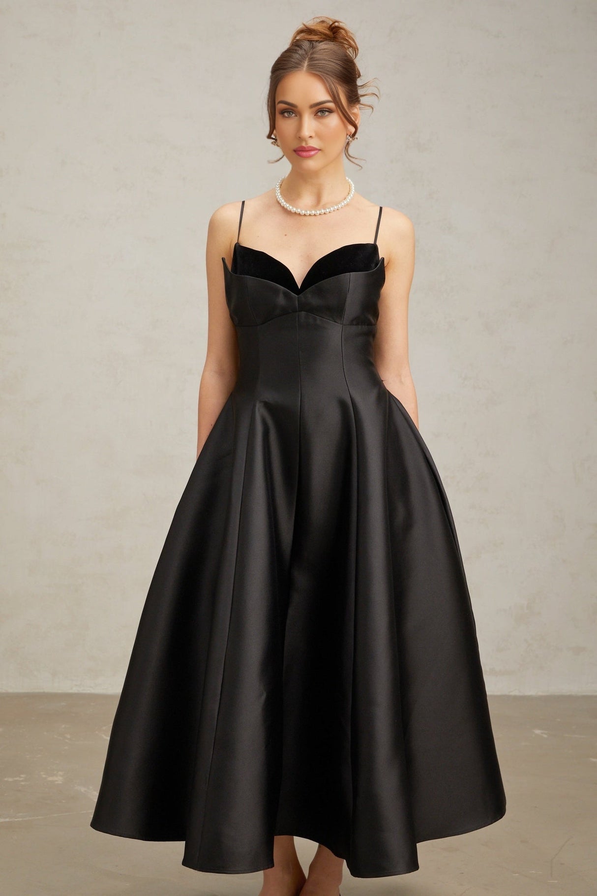 Woman wearing a black pleated midi dress with flared skirt and fitted waist standing against plain background