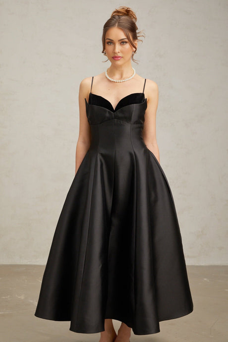 Woman wearing a black pleated midi dress with flared skirt and fitted waist standing against plain background