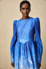 Woman wearing a blue midi dress with gradient color effect and pleated skirt standing against plain background