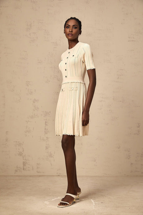 Woman wearing a lightcolored knit mini dress with pleated skirt and delicate pointelle trim detail standing in a neutral setting