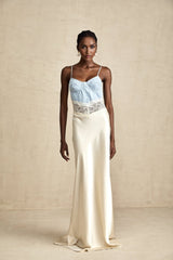 Woman wearing a long flowing maxi dress with lace trim and paneled design in a light color standing against a plain background