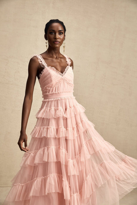 Woman wearing a long flowy layered maxi dress with ruffled tulle skirt in pale color standing against plain background