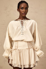 Woman wearing a matching set consisting of a light colored ruffled blouse with self tie details and a coordinating skirt standing against a plain background