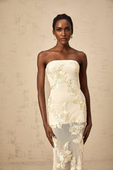 Woman wearing a neutralcolored offshoulder maxi dress with embroidery details standing in a relaxed pose