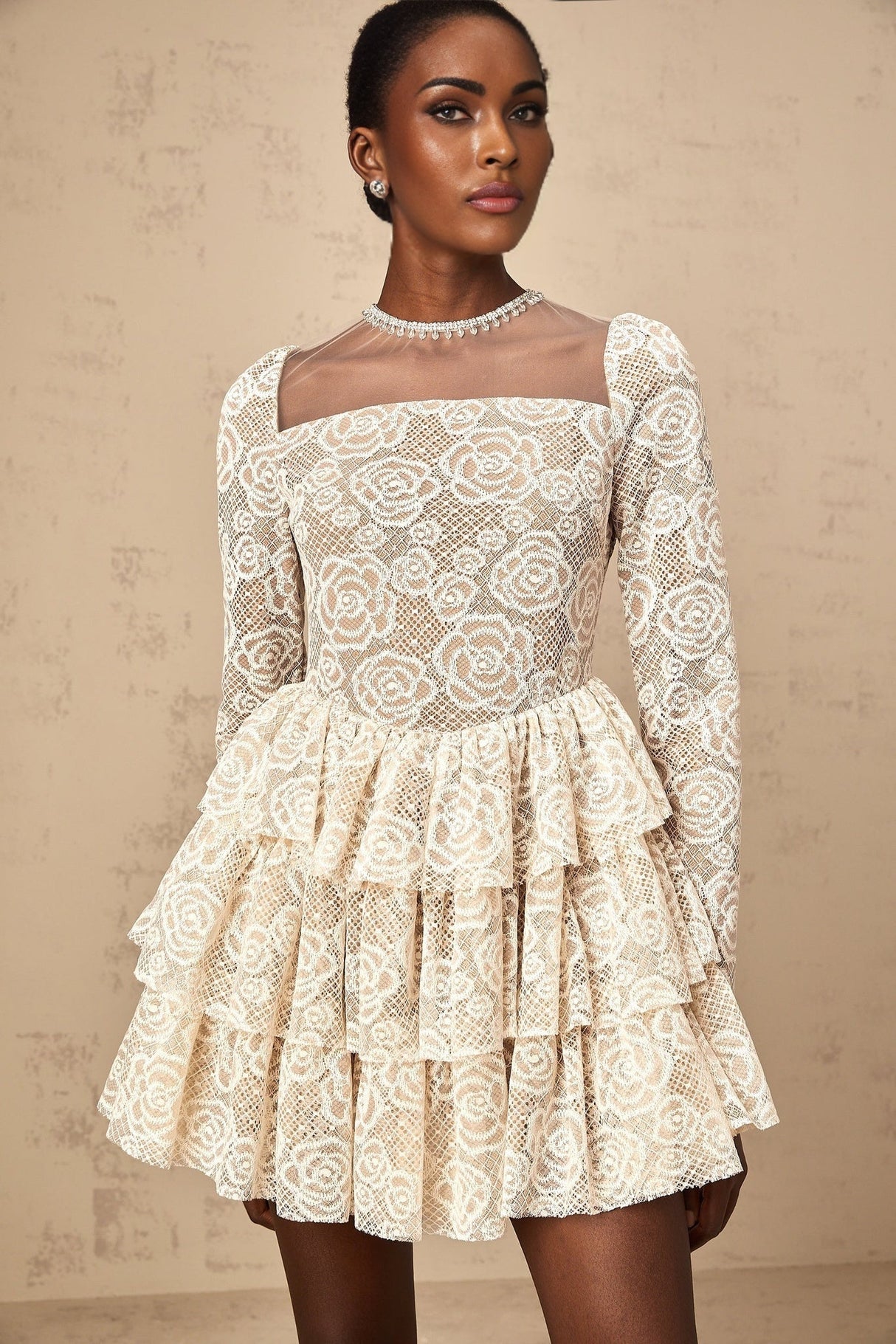 Woman wearing a short beige dress with floral embroidery details standing against a plain background