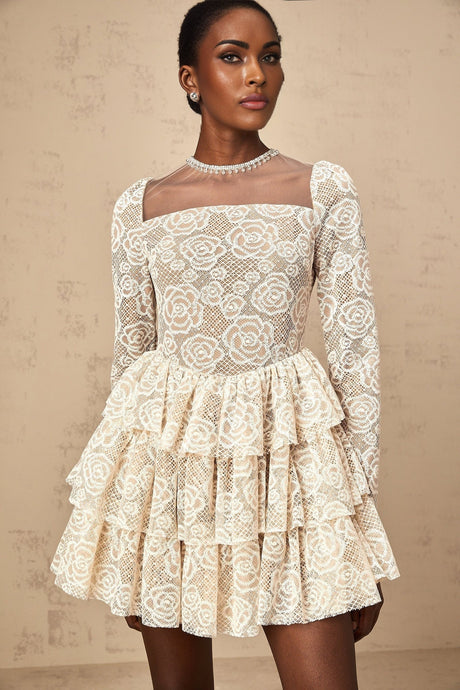 Woman wearing a short beige dress with floral embroidery details standing against a plain background