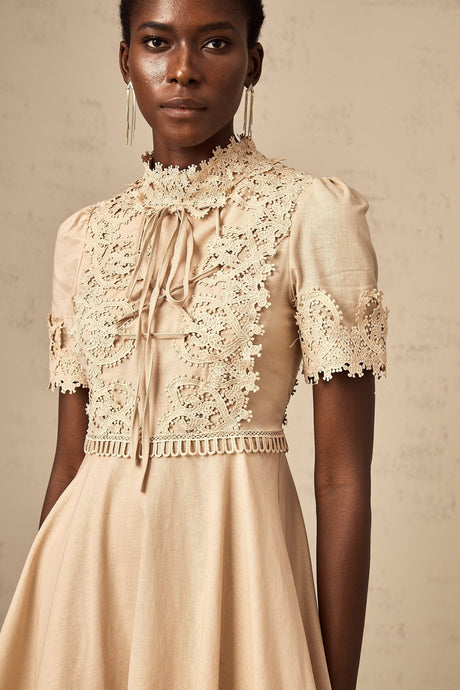 Woman wearing a short flared dress with corded lace pattern in light color standing against plain background