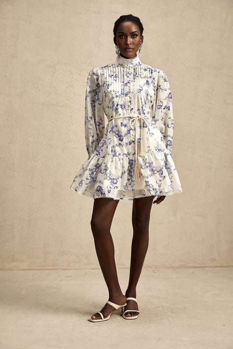 Woman wearing a short floral print dress with flared sleeves and a flowy chiffon fabric in pastel colors standing against a plain background