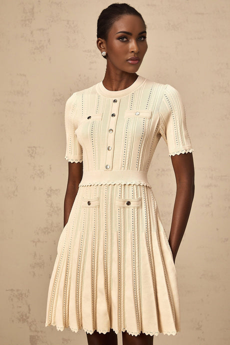 Woman wearing a short neutral colored knit dress with pleated skirt and delicate pointelle trim detail