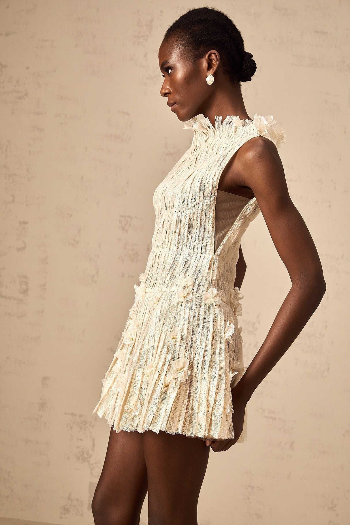 Woman wearing a short white lace dress with semi sheer design and creased texture standing against plain background