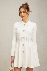 Woman wearing a short white pleated knit dress with long sleeves standing against a plain background