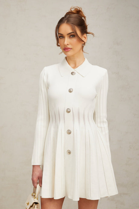 Woman wearing a short white pleated knit dress with long sleeves standing against a plain background
