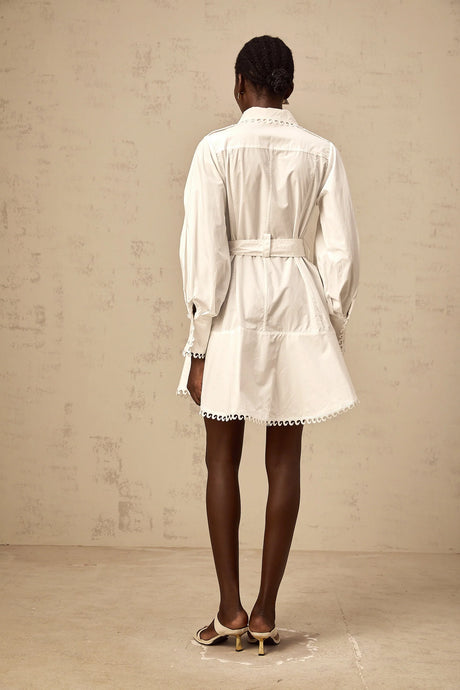Woman wearing a short white shirt dress with belt cinched at waist standing against plain background