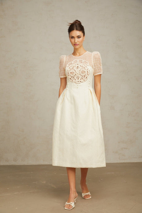 Woman wearing a white midi dress with braided panel details and sleeveless design standing against plain background