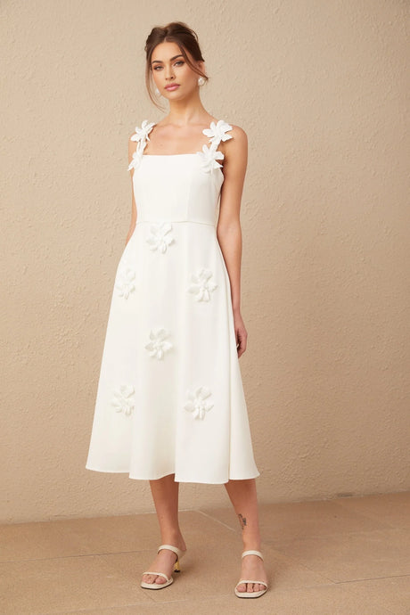 Woman wearing a white midi dress with square neckline and faux flower details standing against plain background