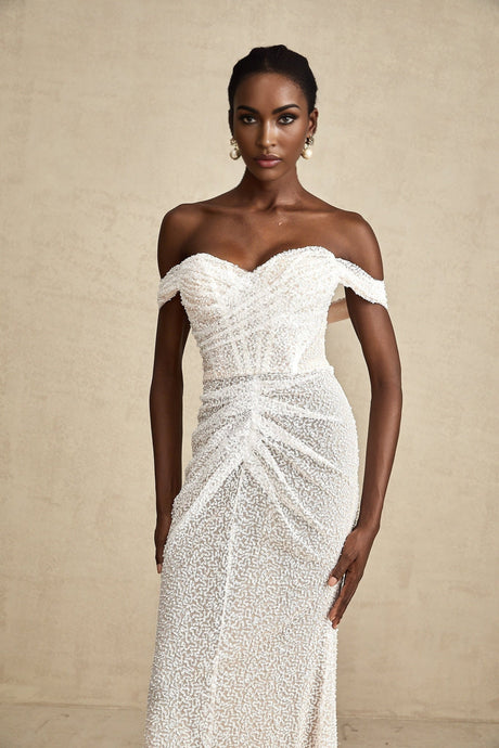 Woman wearing a white offshoulder maxi dress with embellishments standing in a neutral background