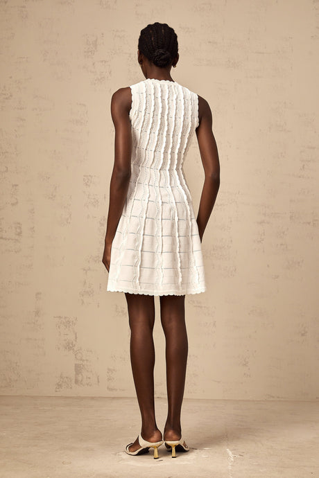 Woman wearing a white sleeveless knit mini dress with ruffled details standing against a plain background