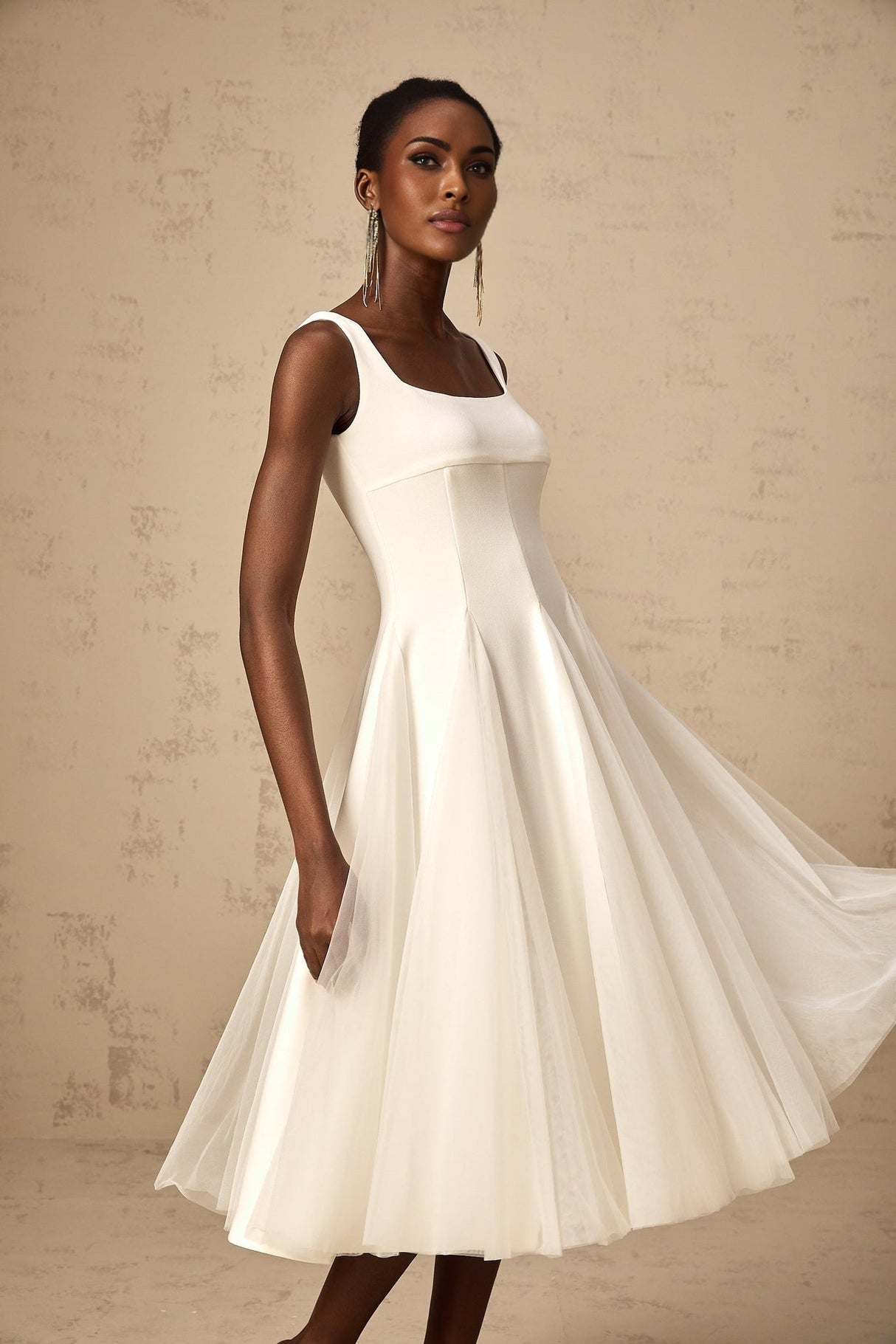Woman wearing a white tulle midi dress with godet inserts and flared skirt standing in a neutral background