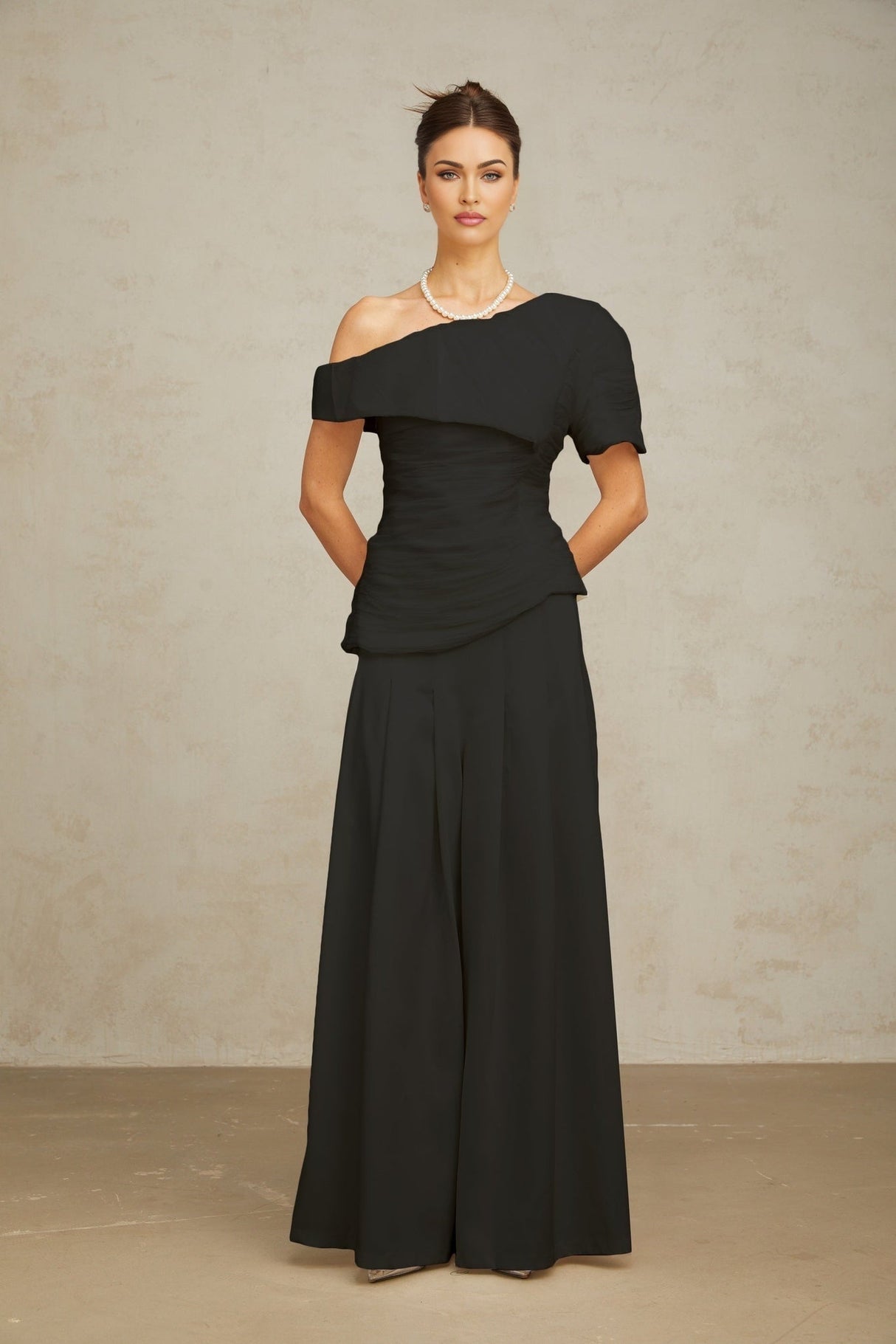 Woman wearing black creased top and matching trousers standing against plain background