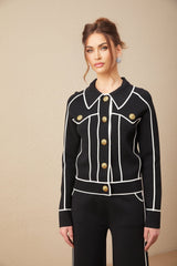 Woman wearing black jacket and trousers set with white piping trim standing against plain background