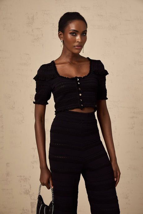 Woman wearing black knitted matching set with ruffled top and trousers standing against plain background