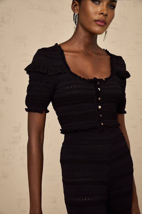 Woman wearing black knitted matching set with ruffled top and trousers standing against plain background