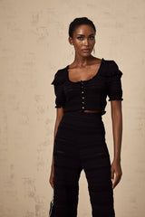 Woman wearing black knitted ruffled top and matching trousers set standing against plain background