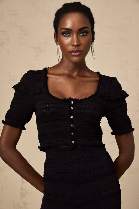 Woman wearing black knitted ruffled top and matching trousers standing against plain background