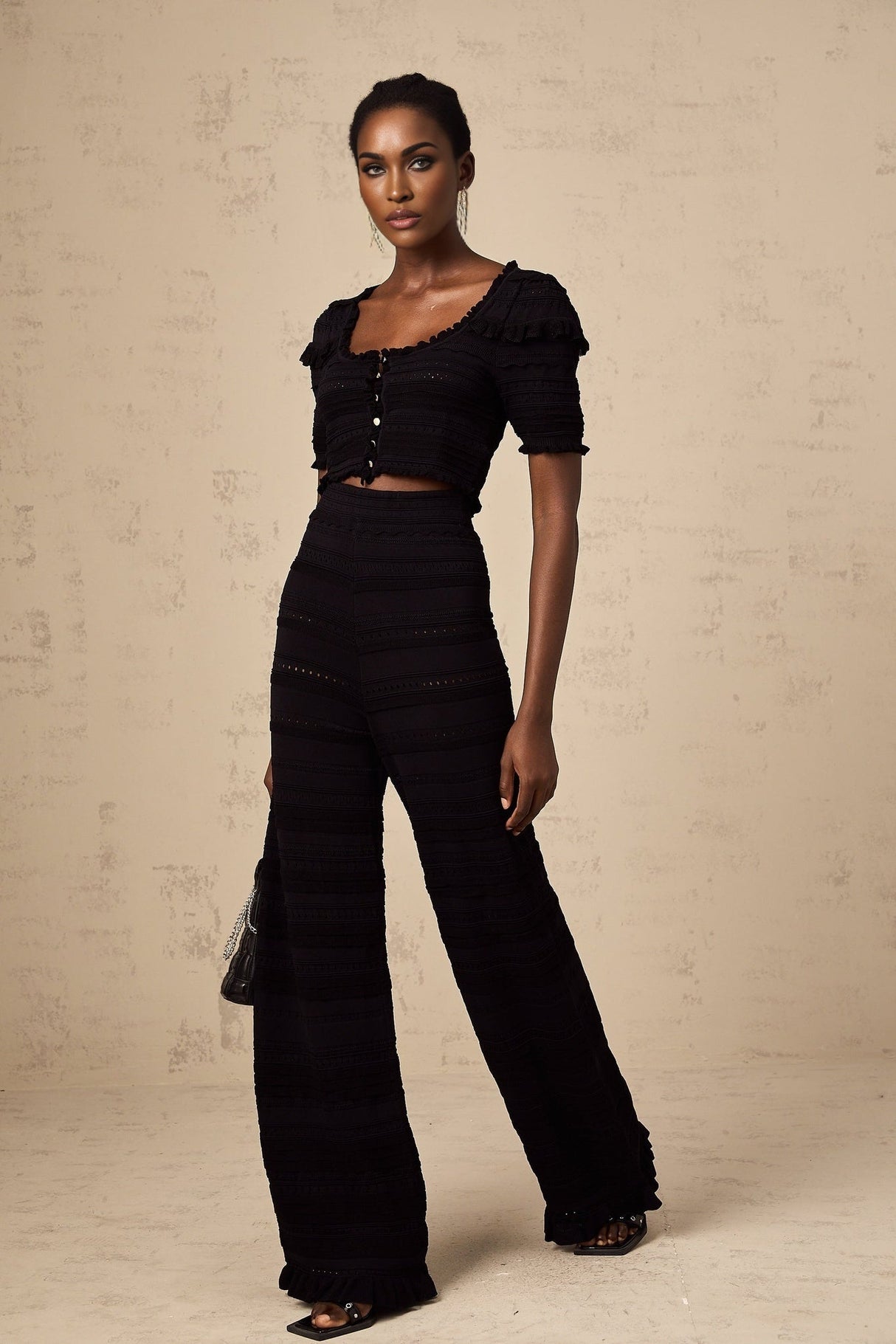 Woman wearing black knitted top with ruffled details and matching trousers standing against plain background
