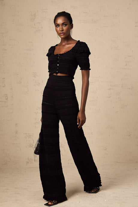 Woman wearing black knitted top with ruffled details and matching trousers standing against plain background