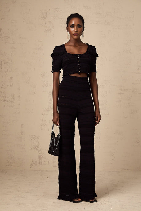 Woman wearing black knitted top with ruffled details and matching trousers standing against plain background