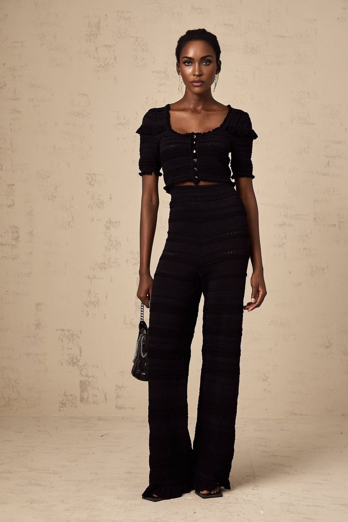 Woman wearing black knitted top with ruffled details and matching trousers standing against plain background