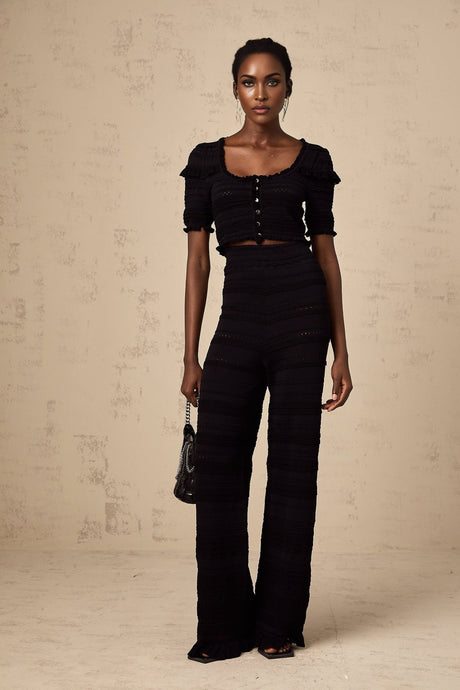 Woman wearing black knitted top with ruffled details and matching trousers standing against plain background