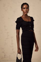 Woman wearing black knitted top with ruffled details and matching trousers standing against plain background