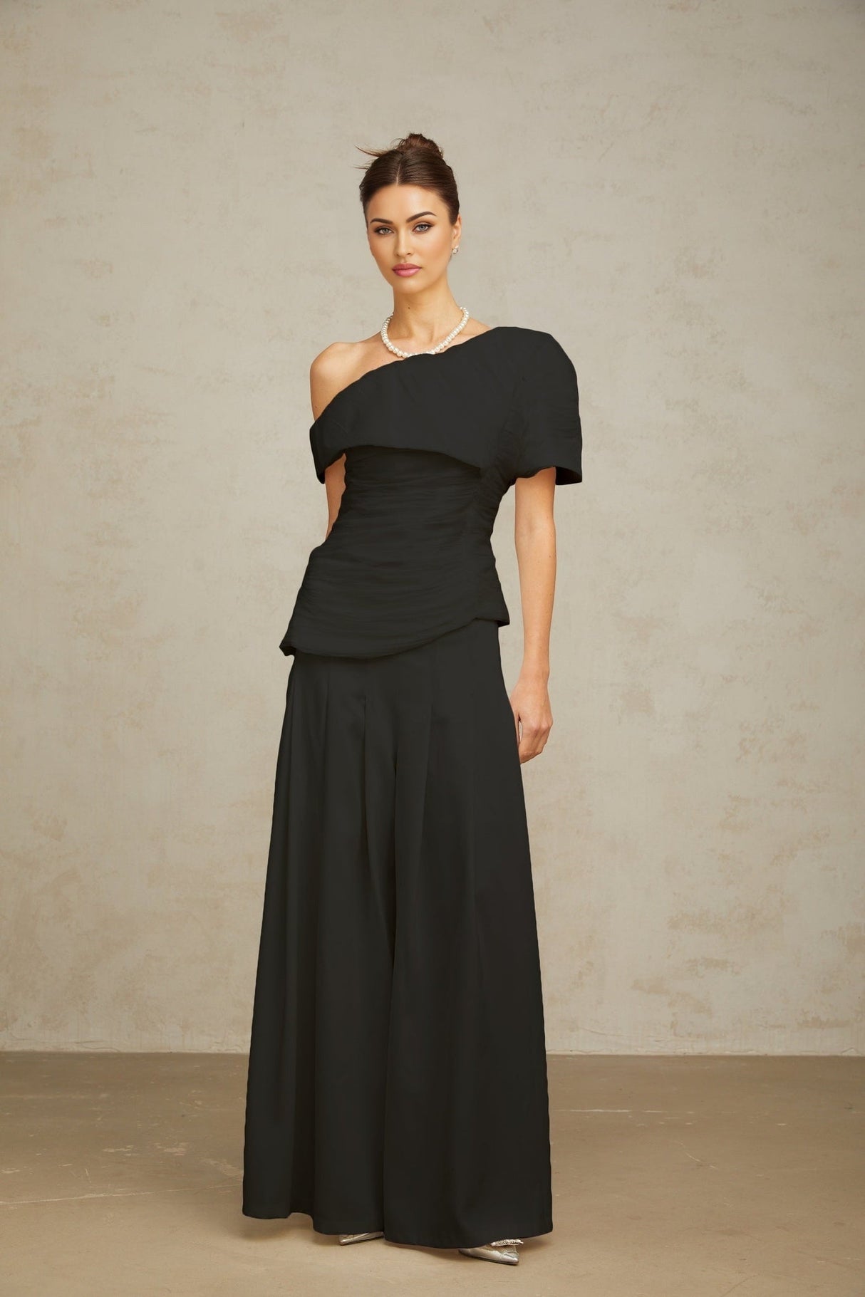 Woman wearing black matching set with creased top and trousers standing against plain background
