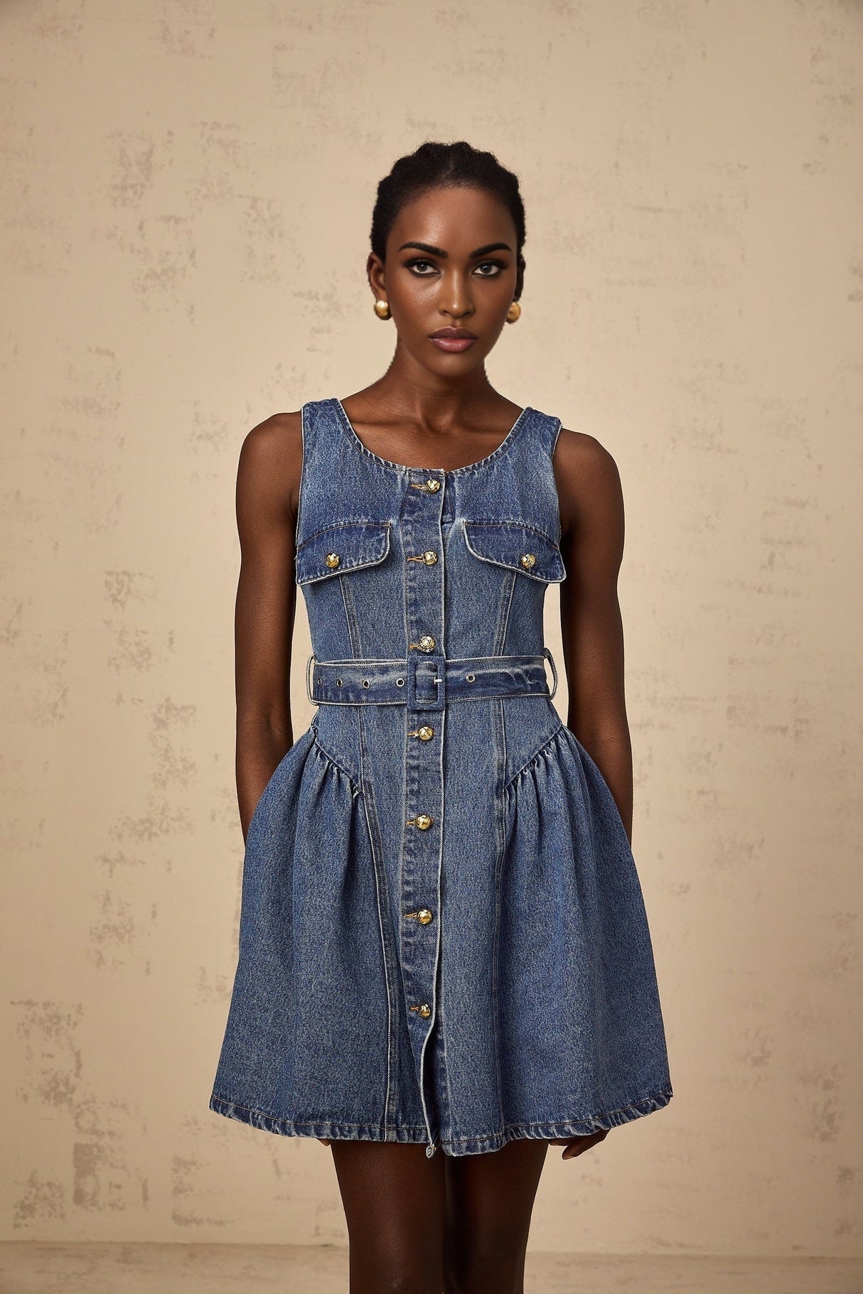 Woman wearing blue denim mini dress with belt cinched at waist standing against plain background