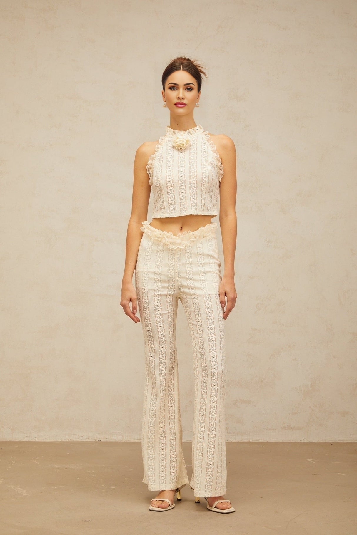 Woman wearing cream white jacquard matching set with ruffled lace trim top and trousers standing against plain background