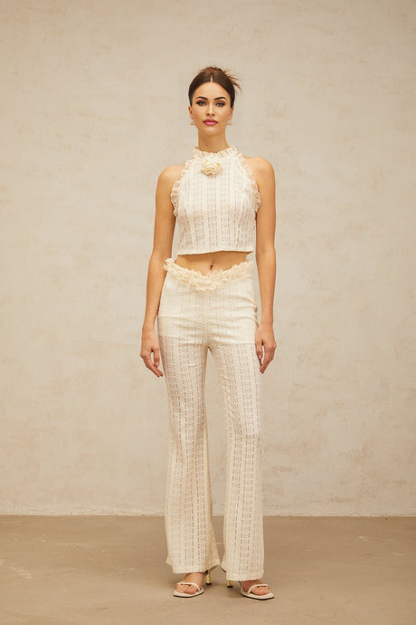Woman wearing cream white jacquard matching set with ruffled lace trim top and trousers standing against plain background