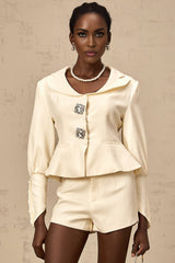 Woman wearing crystal embellished jacket and matching shorts set in white color with sparkly details standing against plain background