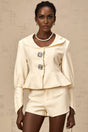 Woman wearing crystal embellished jacket and matching shorts set in white color with sparkly details standing against plain background