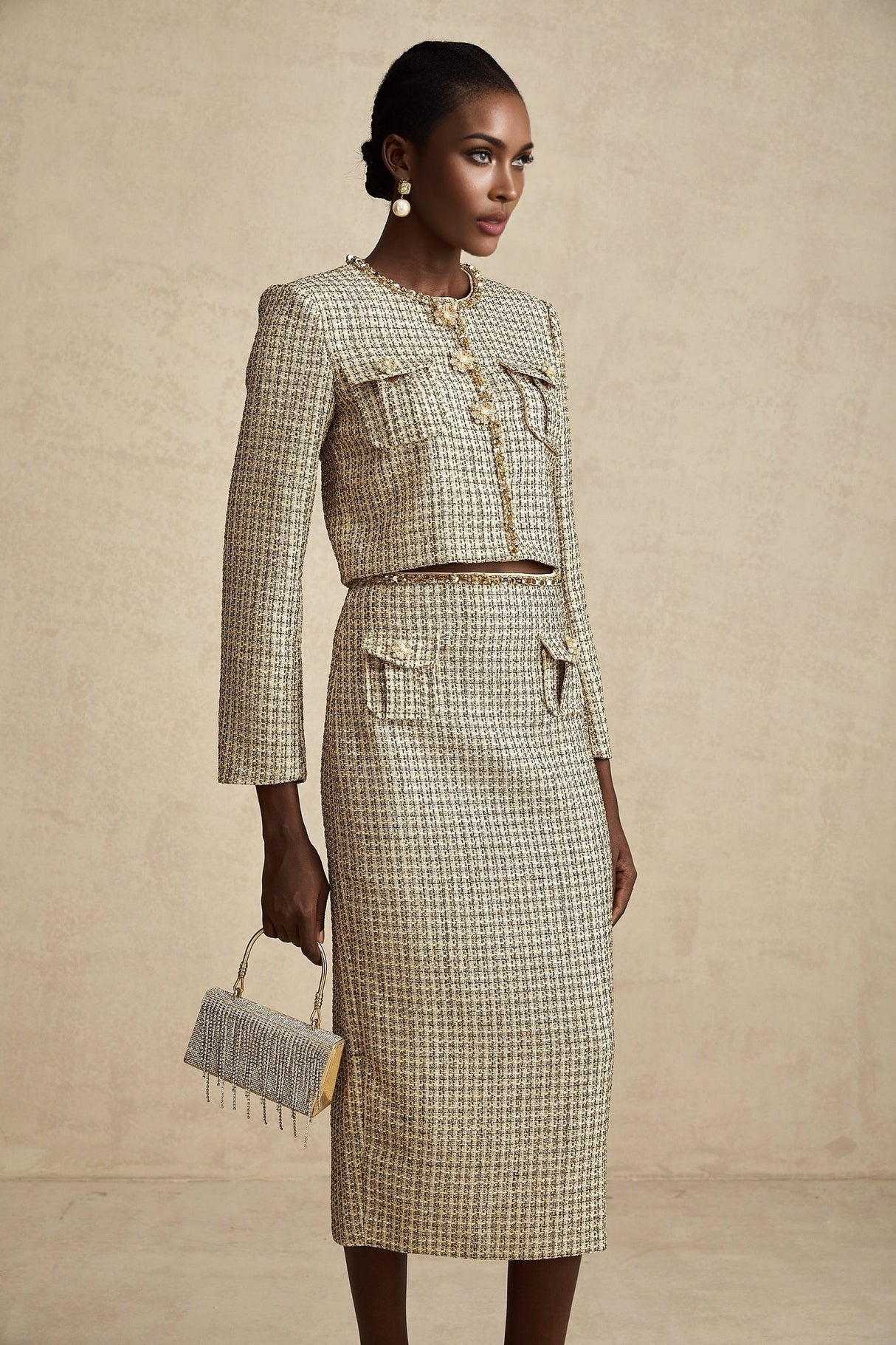 Woman wearing gold tweed sequin jacket and matching skirt set by Aelle standing against plain background